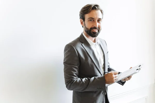 Imagen del exitoso empresario feliz sosteniendo el portapapeles y escribiendo notas mientras trabaja en la oficina — Foto de Stock