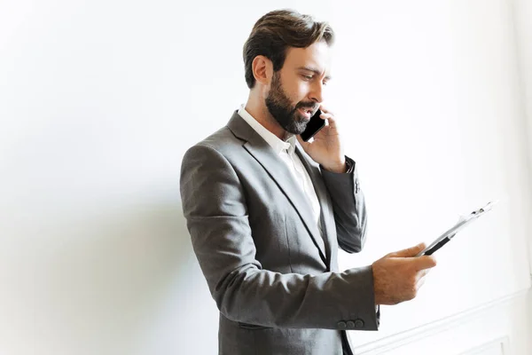 Kuva vakavasta älykkäästä liikemiehestä puhumassa kännykällä ja pitämässä leikepöytää virassa työskennellessään — kuvapankkivalokuva