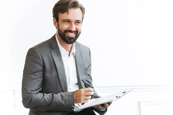 Imagen del hombre de negocios alegre barbudo mirando hacia adelante y sosteniendo el portapapeles mientras trabaja en la oficina —  Fotos de Stock