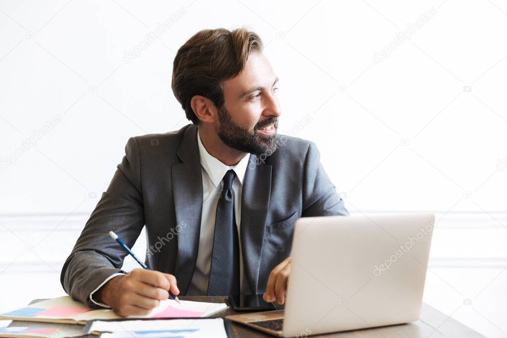 Image of smiling bearded businessman working at laptop in office while writing down notes