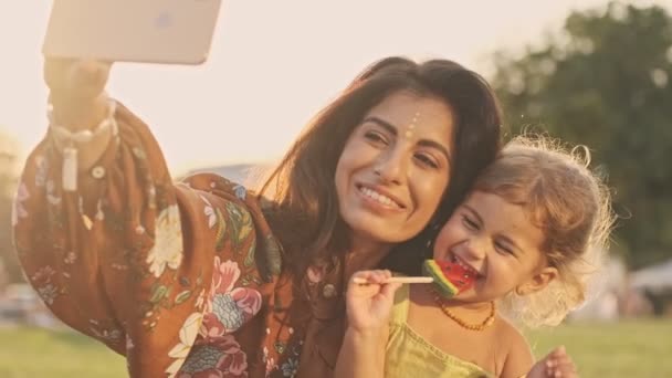 陽気なインド人女性作る自撮り上のスマートフォンとともに彼女の幸せな子供女の子それ食べるロリポップで公園屋外 — ストック動画
