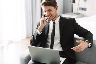 Evde cep telefonuyla konuşan bilgisayarla gülümseyen mutlu genç iş adamı..