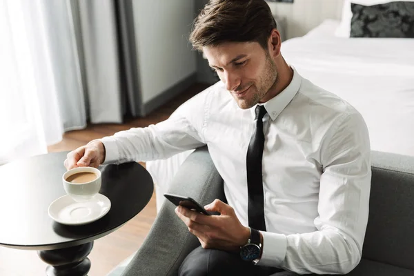 Image of joyful young man holding smartphone and drinking coffee — Stock Photo, Image
