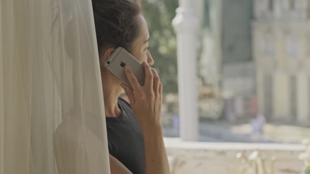 窓の近くに立って 自宅でスマートフォンで話している幸せなアジアの女性の側面図 — ストック動画