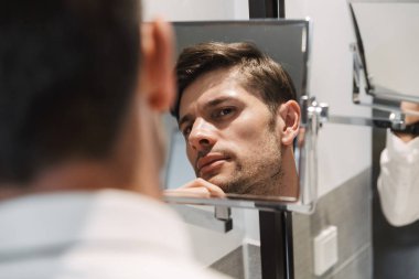 Tıraşsız bir adamın banyodaki aynaya bakışının görüntüsü.