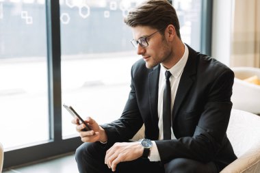 Yakışıklı bir gencin fotoğrafı. Otel odasında akıllı telefonuyla oturuyor.