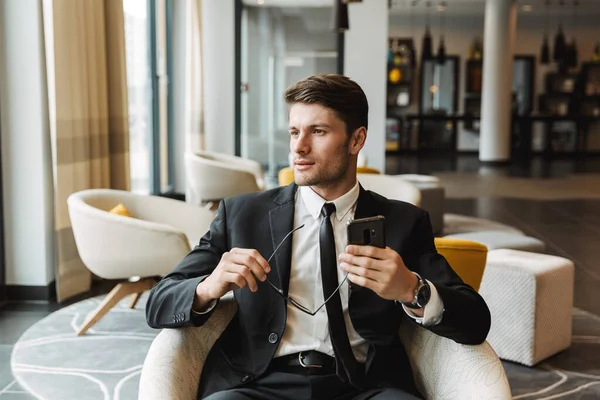 Photo of attractive young man sitting in hotel hall with smartph — Stock Photo, Image