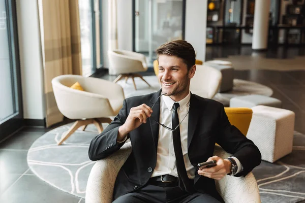 Photo of joyful young man sitting in hotel hall with smartphone — Stock Photo, Image