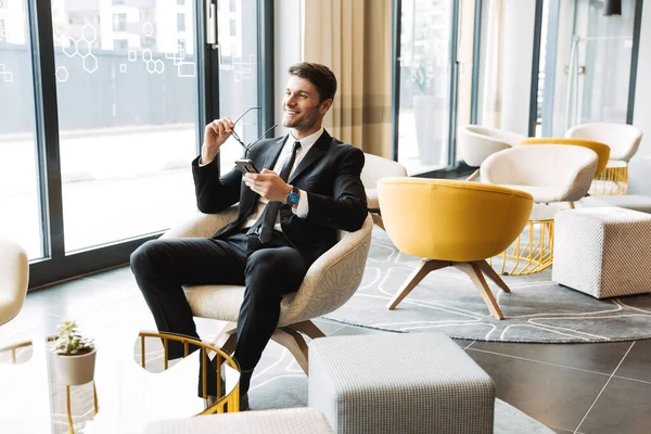 Photo of smiling young man sitting in hotel hall with smartphone — Stock Photo, Image