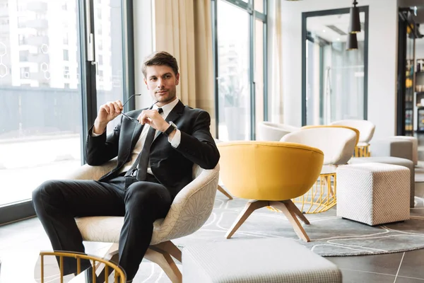 Photo of confident young man holding eyeglasses in hotel hall du — Stock Photo, Image