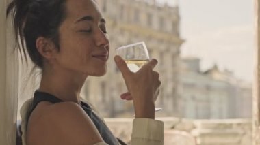 Keyifli, kaygısız, güzel Asyalı kadın şarap içerken ve bu anın tadını çıkarırken evinin penceresinin yanındaki koltukta otururken.