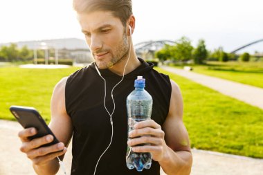 Green Park 'ta kulaklıkla koşarken su içen ve cep telefonu tutan yakışıklı bir adam.