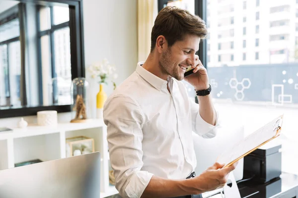 Imagen del exitoso hombre de negocios sosteniendo portapapeles con papeles y hablando por teléfono celular mientras trabaja en la oficina —  Fotos de Stock