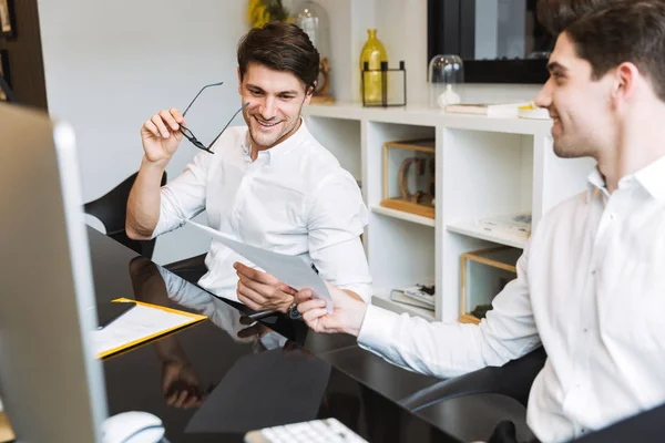 Изображение двух счастливых мужчин с бумажными документами, сидящих за столом и работающих за компьютерами в офисе — стоковое фото