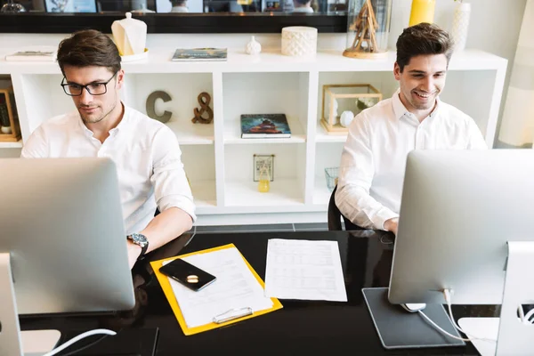 Изображение двух улыбающихся мужчин, сидящих за столом и работающих за компьютером — стоковое фото