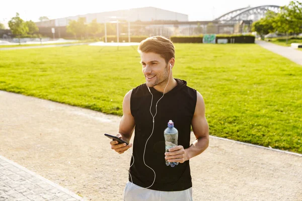在室外绿地公园，肌肉发达的男子一边喝水，一边拿着手机，一边拿着耳机跑步的照片 — 图库照片