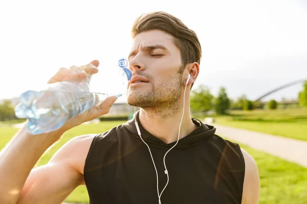 Изображение кавказца, пьющего воду во время тренировки с наушниками и бегущего в зеленом парке утром — стоковое фото