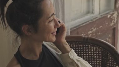 Neşeli, kaygısız, güzel Asyalı kadının akıllı telefonuyla konuşması ve evinin penceresinin yanındaki koltukta otururken gözlerini kaçırması.