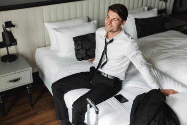 Otelde bavulla yatakta oturan memnun genç bir adam resmi.