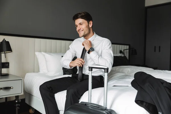 Imagem do jovem feliz sentado na cama com mala no hotel r — Fotografia de Stock