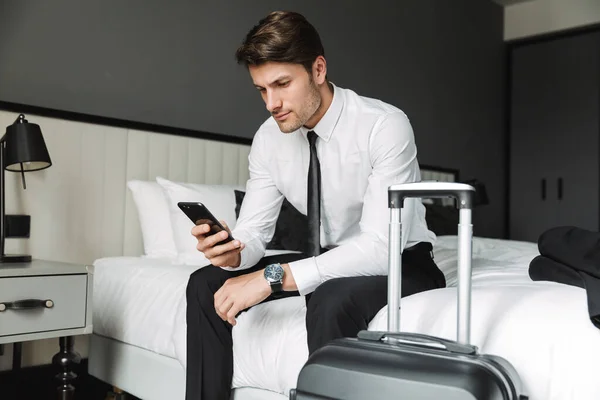 Imagem do jovem moderno segurando celular enquanto sentado na cama — Fotografia de Stock