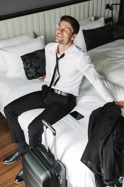 笑顔の若い男がベッドの上でスーツケースと一緒にホテルに座っているイメージ — ストック写真