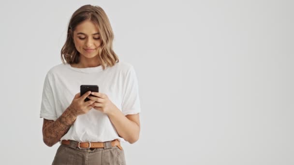 漂亮的年轻女人穿着一件白色的基本T恤 一边用智能手机 一边看着被白色背景隔开的侧面 — 图库视频影像