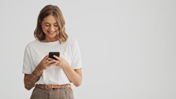 Uśmiechnięta Młoda Kobieta Nosi Podstawowy Shirt Pomocą Smartfona Wskazując Bok — Wideo stockowe