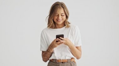 Beyaz arka planda izole edilmiş akıllı telefonunu kullanarak beyaz tişört giyen çekici genç bir kadın.