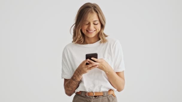 迷人的年轻女子穿着白色的基本T恤 用她的智能手机与白色背景隔离 — 图库视频影像