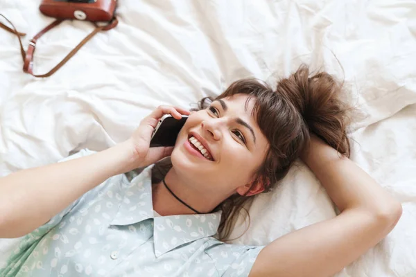Young positive smiling pretty woman indoors at home lies on bed talking by mobile phone. — Stock Photo, Image