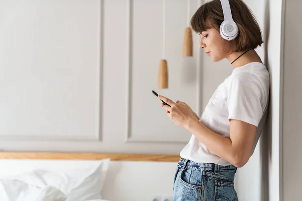 집에서 헤드폰을 끼고 핸드폰을 사용하여 음악을 듣고 있는 놀라운 아가씨. — 스톡 사진