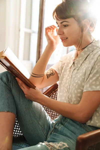 Όμορφη γυναίκα μέσα στο σπίτι κάθεται σε καρέκλα διαβάζοντας το βιβλίο. — Φωτογραφία Αρχείου