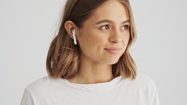 年轻漂亮的女人穿着一件白色的基本T恤 开始在她的无线耳机里听音乐 这些耳机是白色背景的 — 图库视频影像