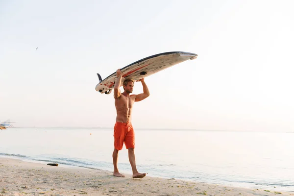 Imagen del hombre surfista musculoso llevando su tabla de surf por el océano en —  Fotos de Stock