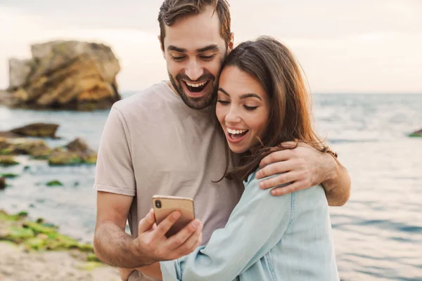 Fénykép izgatott fiatal pár segítségével mobiltelefon és ölelés — Stock Fotó