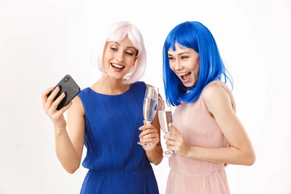 Retrato de duas mulheres sorridentes vestindo perucas azuis e rosa tirando foto selfie no celular enquanto bebe champanhe — Fotografia de Stock