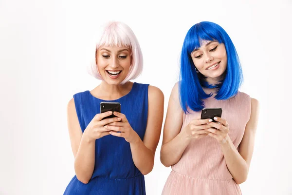 Két boldog fiatal nő portréja, akik kék és rózsaszín parókát viselnek, mosolyognak, miközben mobilon gépelnek. — Stock Fotó