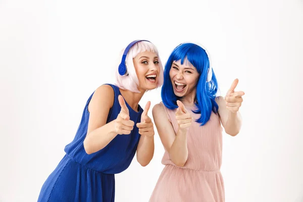 Porträt zweier fröhlicher Frauen mit blauen und rosa Perücken, die mit dem Finger auf die Kamera zeigen, während sie Kopfhörer benutzen — Stockfoto