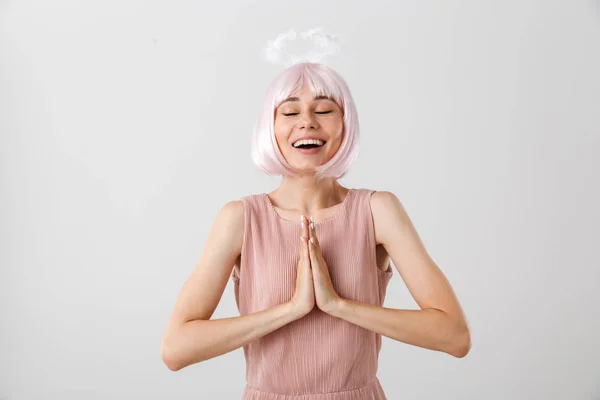 Portret uśmiechniętej miłej kobiety ubranej w różową perukę i zabawkę halo marzącej trzymając dłonie razem — Zdjęcie stockowe