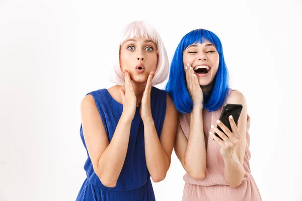 Portret dwóch radosnych kobiet noszących niebieskie i różowe peruki robiących śmieszne miny podczas korzystania z telefonu komórkowego — Zdjęcie stockowe