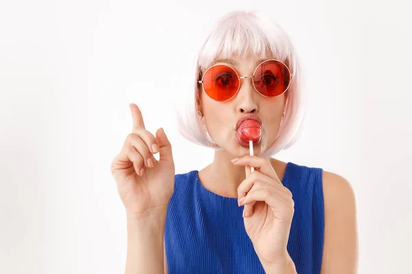 Portret van een jonge blanke vrouw met een roze pruik en een bril die lolly eet terwijl ze met haar vinger naar boven wijst — Stockfoto