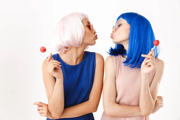 Portret van twee jonge grappige vrouwen met pruiken en brillen die lolly 's vasthouden terwijl ze hun gezicht kussen — Stockfoto