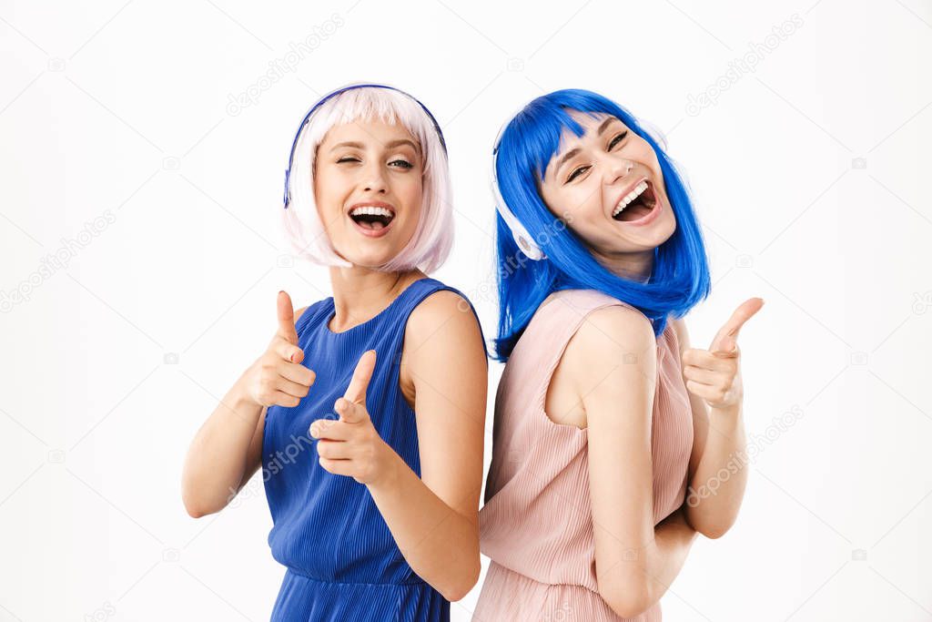 Portrait of two laughing women wearing blue and pink wigs pointing fingers at camera while using headphones