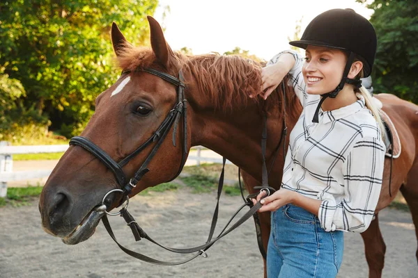 Εικόνα της ξανθιάς γυναίκας φορώντας καπέλο στέκεται από άλογο στην αυλή σε γ — Φωτογραφία Αρχείου