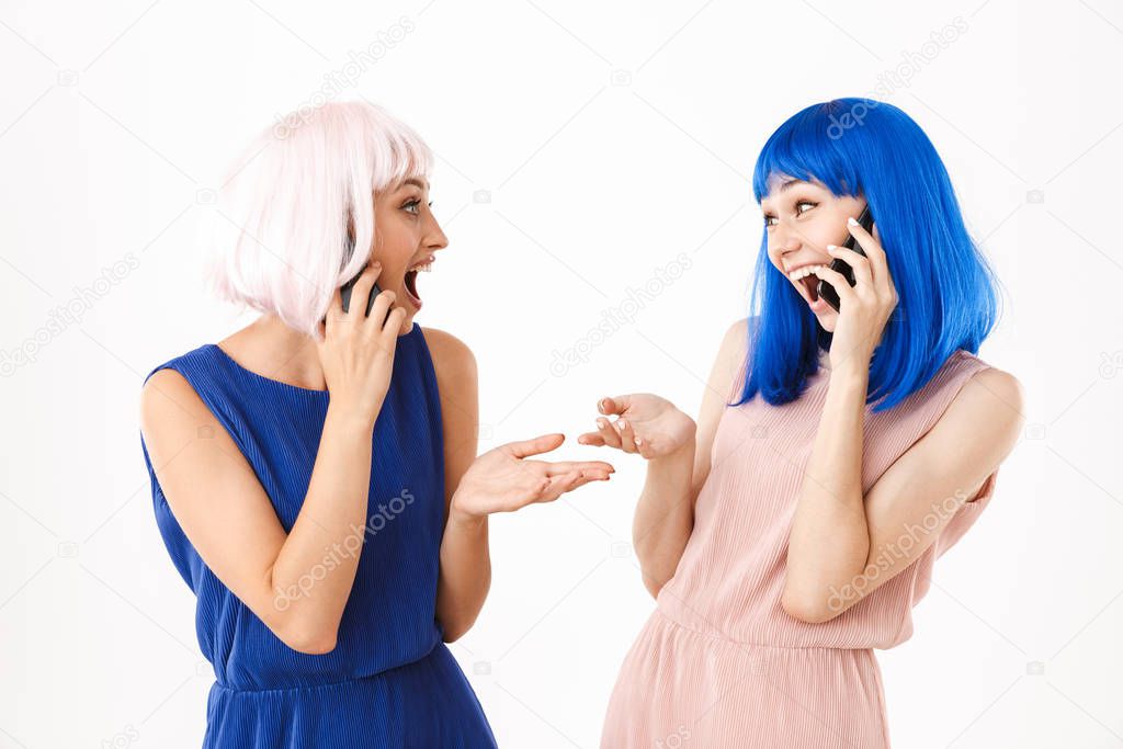 Portrait of two delighted women wearing blue and pink wigs looking at each other while talking on cellphones