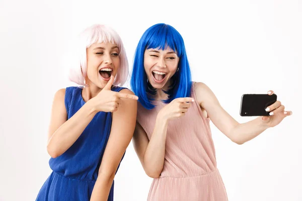 Két vicces, kék és rózsaszín parókát viselő nő portréja, akik szelfi fotókat készítenek és ujjal mutogatnak a mobiltelefonra. — Stock Fotó