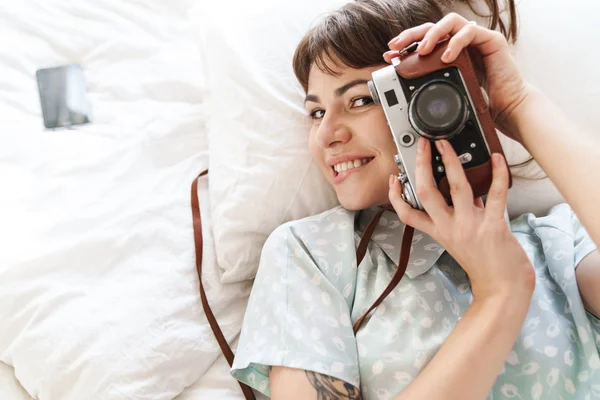 Gelukkig vrolijke positieve vrouw binnen ligt op bed in de ochtend met camera fotograferen. — Stockfoto