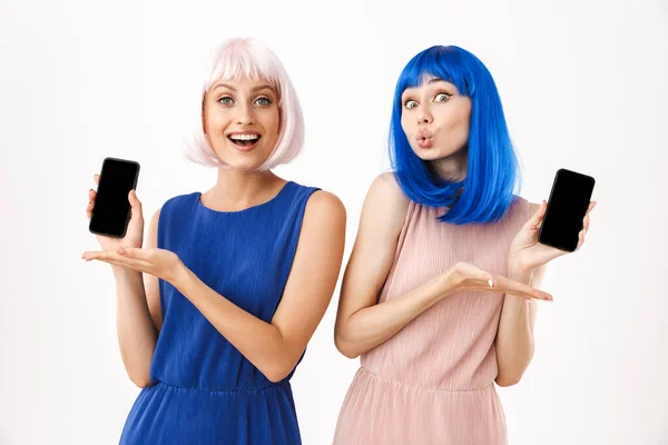 Két örömteli nő portréja, kék és rózsaszín parókát viselnek, vicces arcot vágnak, miközben mobiltelefonokat tartanak a kezükben. — Stock Fotó