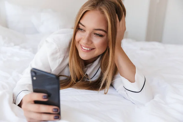 Foto de mujer alegre agradable usando el teléfono celular y sonriendo mientras que miente —  Fotos de Stock